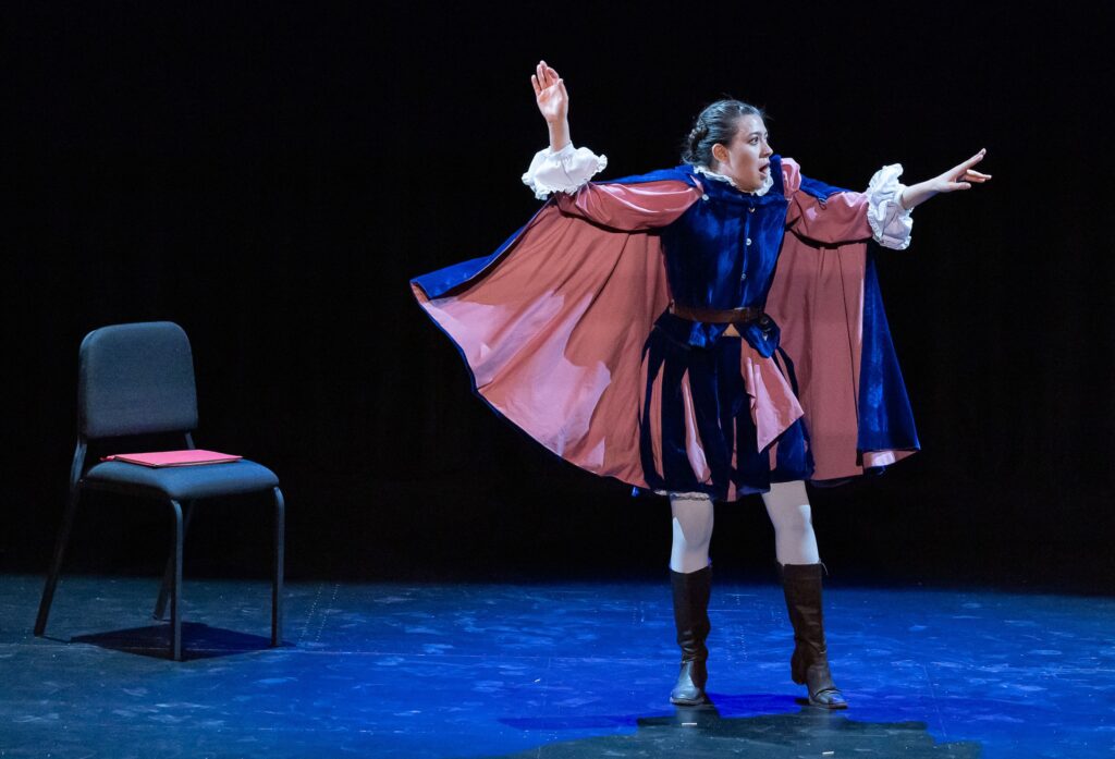 Woman on stage in a cape and breeches, with arms raised and pointing right