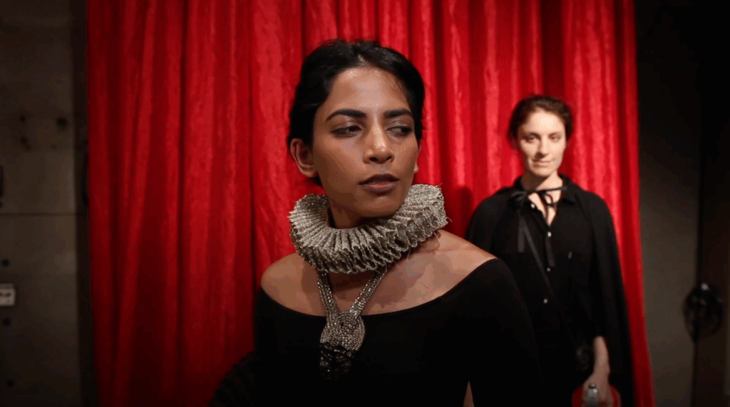 Click here to watch scenes. The image is of a woman glancing over her shoulder to a another woman standing behind her. 